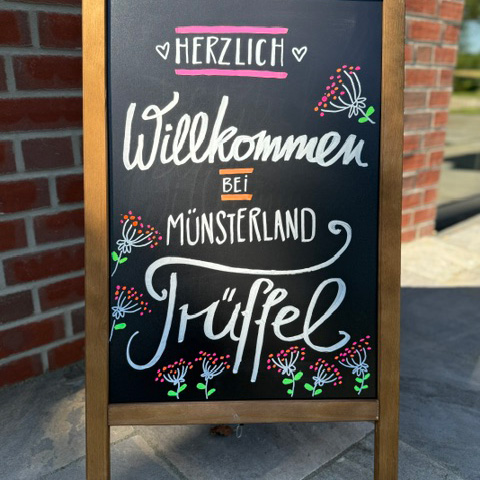 Eine Tafel mit Lettering und floralen Elementen. Der Text lautet: Herzlich Willkommen bei Münstarlandtrüffel.